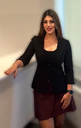 Aynur Dönmez, siyah ceket ve bordo etekle, beyaz bir tezgâha elini dayamış gülümsüyor